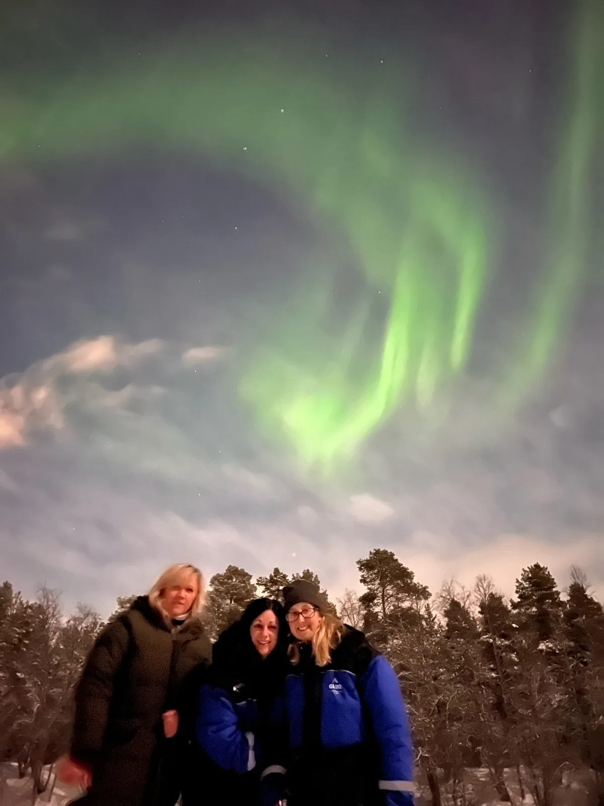 Three guests in GLØD winter suits smiling under a green aurora swirl above snowy pines
